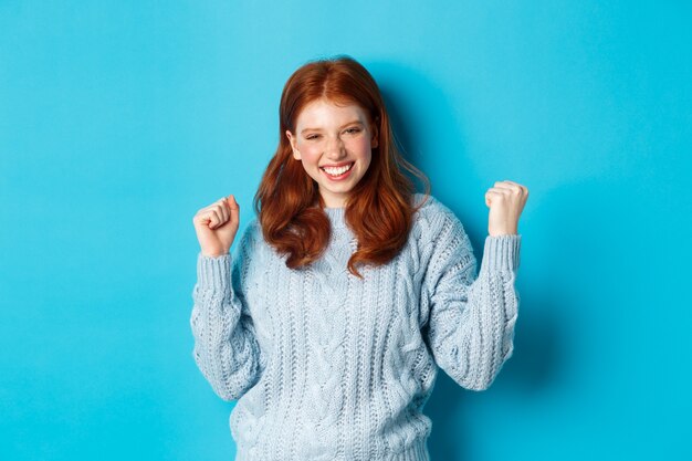 La ragazza rossa soddisfatta raggiunge l'obiettivo e festeggia, facendo il gesto della pompa del pugno e sorridendo felice, trionfando della vittoria, in piedi su sfondo blu