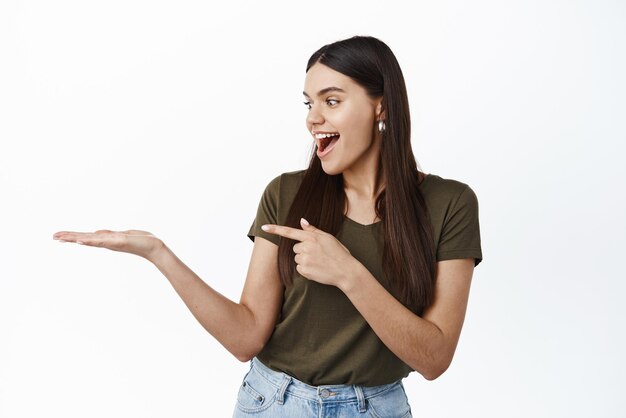 La ragazza sembra eccitata al suo palmo con l'annuncio dell'elemento che punta a portata di mano contro lo spazio della copia in piedi su sfondo bianco Copia spazio