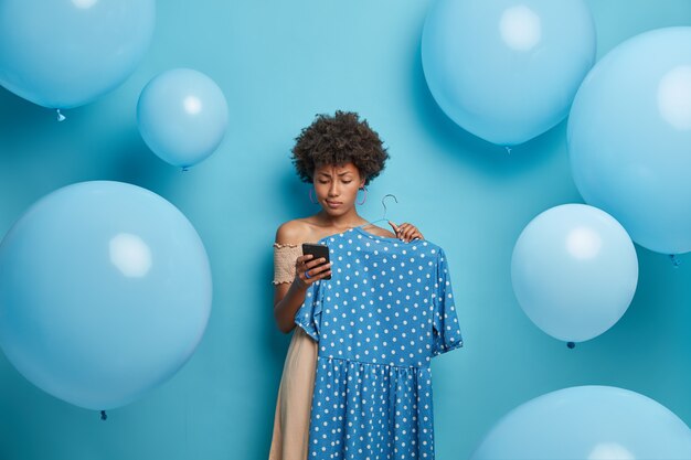 La ragazza seria del compleanno riceve le congratulazioni sullo smartphone, sceglie il vestito blu a pois sulla gruccia, si veste e aspetta gli ospiti, si leva in piedi contro il muro decorato. Donne, vestiti, vestirsi