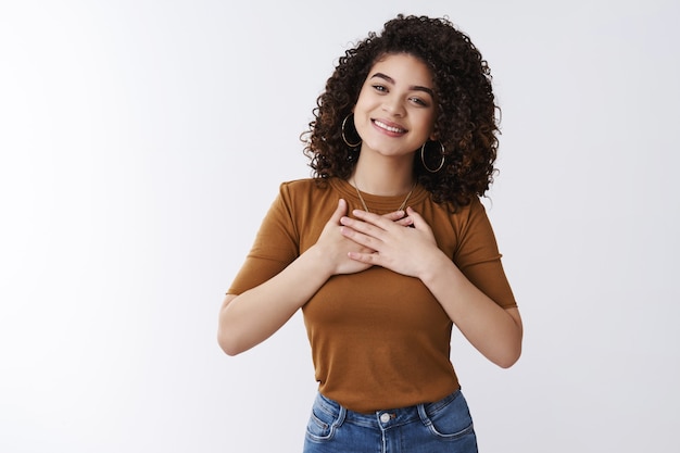 La ragazza si sente grata grazie al cuore in basso. Attraente allegra e moderna giovane donna dai capelli ricci tenere le mani sul petto grata sorridendo compiaciuta come toccare un regalo carino in piedi sfondo bianco apprezzato