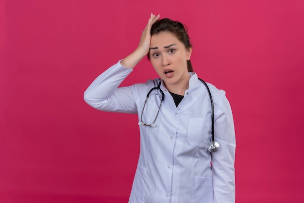 La ragazza sorpresa del medico che porta l'abito medico e lo stetoscopio ha messo la sua mano sulla fronte su priorità bassa rossa parassita