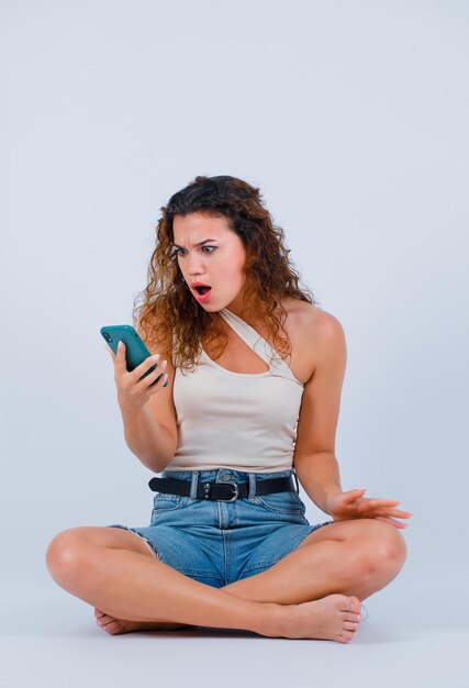 La ragazza sorpresa sta guardando il suo telefono sedendosi su sfondo bianco