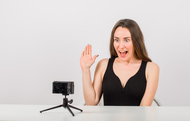 La ragazza sorpresa sta posando alla piccola macchina fotografica mostrando ciao gesto su fondo bianco