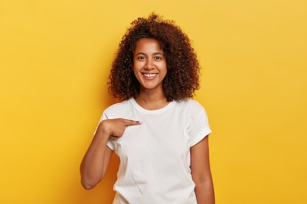 La ragazza sorridente allegra dalla pelle scura indica se stessa, mostra lo spazio del mockup su una maglietta bianca, felice di essere scelta, modelli contro il muro giallo. La giovane donna afro e spensierata mi chiede chi sono