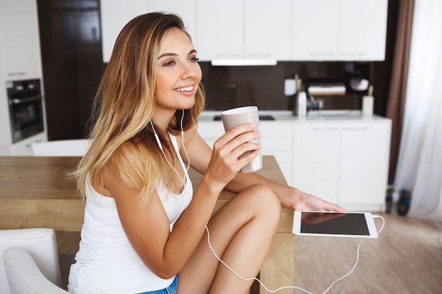 La ragazza sorridente attraente che si siede alla cucina e ascolta la musica.