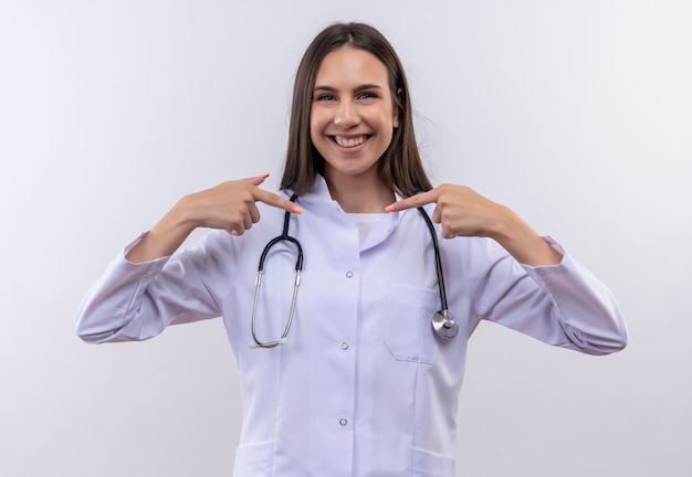 La ragazza sorridente che indossa l'abito medico dello stetoscopio indica se stessa su fondo bianco isolato