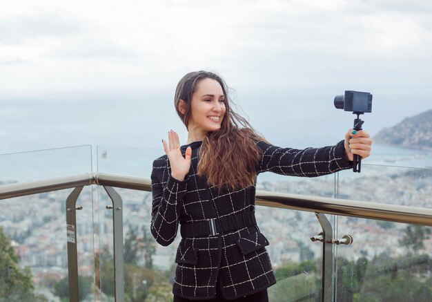 La ragazza sorridente del blogger sta facendo un selfie mostrando un gesto ciao sullo sfondo della vista della città