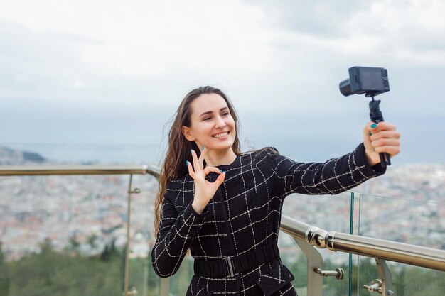 La ragazza sorridente del blogger sta facendo un selfie mostrando un gesto ok alla fotocamera in mano sullo sfondo della vista della città