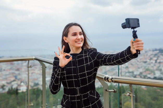 La ragazza sorridente del blogger sta prendendo selfie con la sua mini fotocamera mostrando un gesto perfetto sullo sfondo della vista sulla città