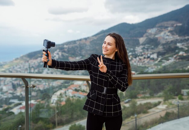 La ragazza sorridente del blogger sta prendendo selfie mostrando il gesto di vittoria sullo sfondo della vista sulla città