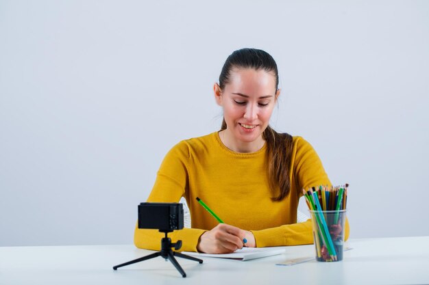 La ragazza sorridente del blogger sta scrivendo un testo sedendosi davanti alla sua mini macchina fotografica su fondo bianco