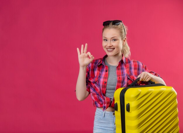 La ragazza sorridente del viaggiatore che porta la valigia della tenuta della camicia rossa mostra il gesto di oker su fondo rosa isolato