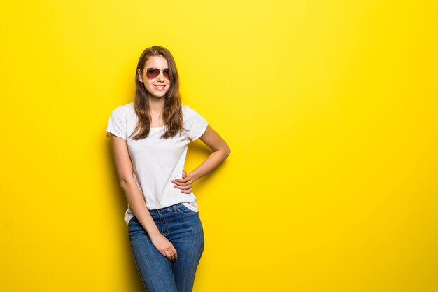 La ragazza sorridente di modo in maglietta bianca e jeans blu rimane davanti al fondo giallo dello studio