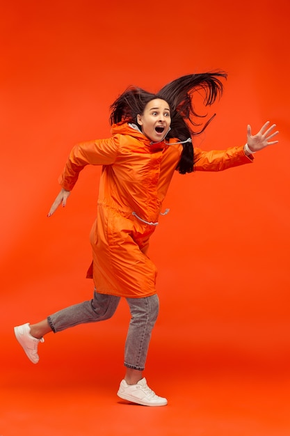 La ragazza sorridente felice che propone allo studio in giacca arancione di autunno isolata sul rosso.