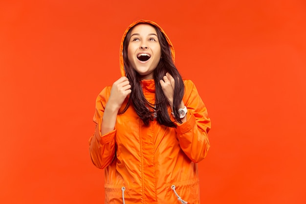 La ragazza sorridente felice che propone allo studio in giacca arancione di autunno isolata sulla parete arancione