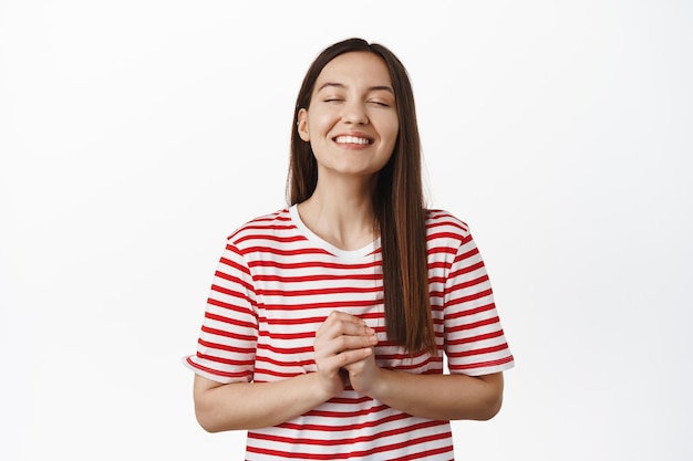 La ragazza sorridente speranzosa chiude gli occhi, tenendosi per mano, pregando o esprimendo desideri, aspettando che accada qualcosa, sognando ad occhi aperti, immagini in bolla sopra la testa, sfondo bianco