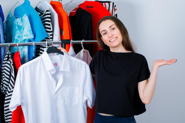 La ragazza sorridente sta esaminando la macchina fotografica tenendo la camicia sullo sfondo dei vestiti