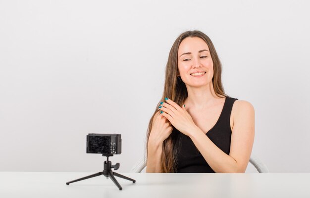 La ragazza sorridente sta esaminando la piccola macchina fotografica tenendo i suoi capelli su fondo bianco