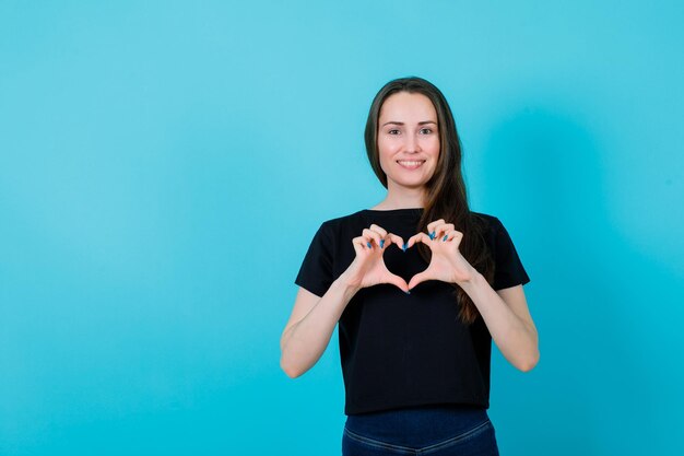 La ragazza sorridente sta mostrando il gesto del cuore collegando le dita su sfondo blu