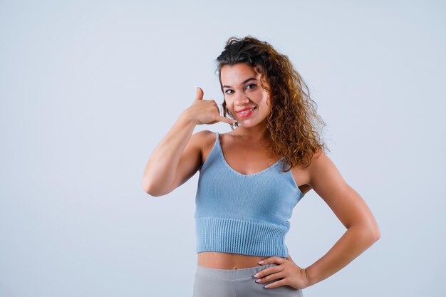 La ragazza sorridente sta mostrando il gesto del telefono e sta mettendo l'altra mano sulla vita su fondo bianco