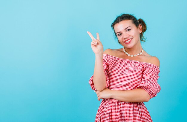 La ragazza sorridente sta mostrando il gesto di vittoria alla fotocamera su sfondo blu