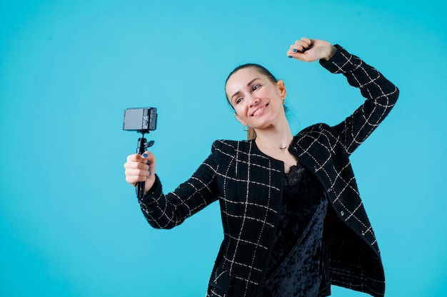 La ragazza sorridente sta prendendo selfie con la sua macchina fotografica alzando il pugno su sfondo blu