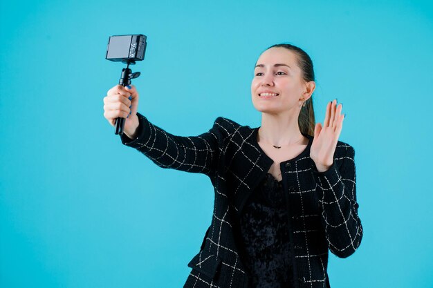 La ragazza sorridente sta prendendo selfie con la sua macchina fotografica e mostra ciao gesto su sfondo blu