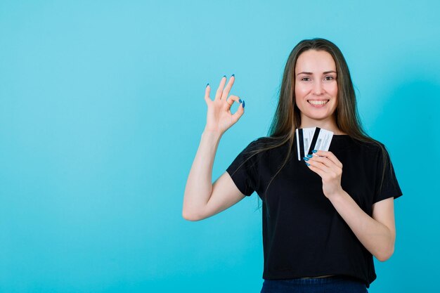 La ragazza sorridente sta tenendo le carte di credito e sta mostrando il gesto giusto su fondo blu