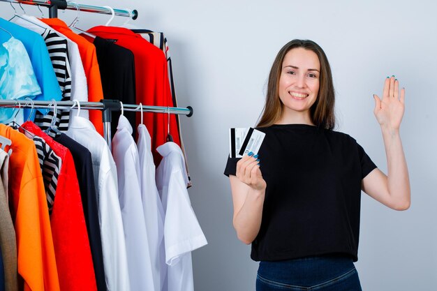 La ragazza sorridente tiene in mano le carte di credito e mostra ciao gesto con l'altra mano sullo sfondo dei vestiti