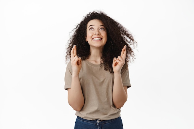 La ragazza speranzosa fa il desiderio di incrociare le dita per buona fortuna e guarda in alto con un sorriso felice anticipando il miracolo pregando Dio affinché il sogno diventi realtà in piedi su sfondo bianco