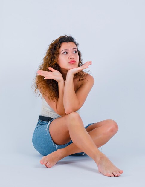 La ragazza sta tenendo le mani sotto il mento sedendosi sul pavimento su fondo bianco