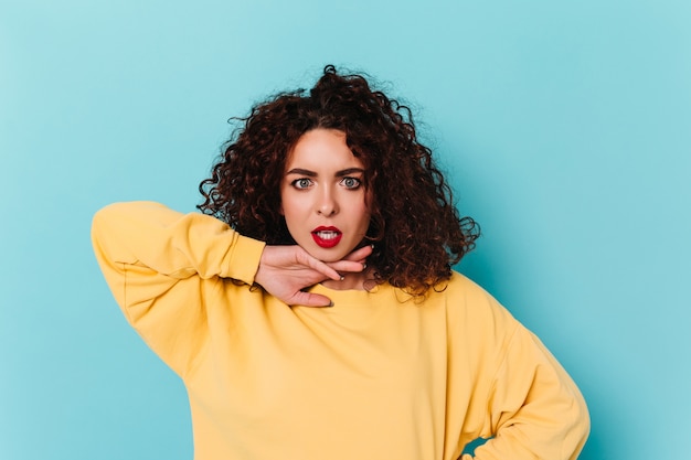 La ragazza stupita con gli occhi azzurri esamina la macchina fotografica, aprendo la bocca. signora con rossetto rosso e capelli scuri ricci sullo spazio blu.