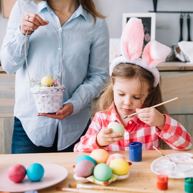 La ragazza sveglia che dipinge le uova per Pasqua vicino alla madre con il piccolo canestro