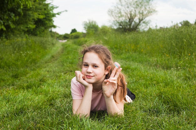 La ragazza sveglia che si trova sulla terra erbosa che mostra la vittoria firma dentro la bella natura