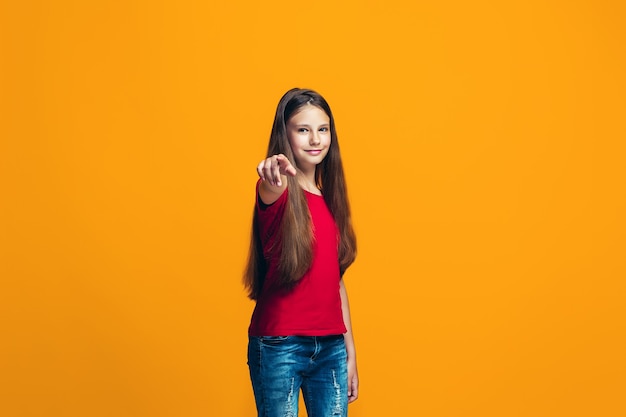 La ragazza teenager felice che indica a voi, ritratto del primo piano di mezza lunghezza su priorità bassa arancione.