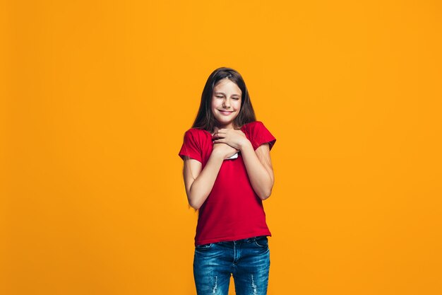 La ragazza teenager felice che sta e che sorride contro la parete arancio