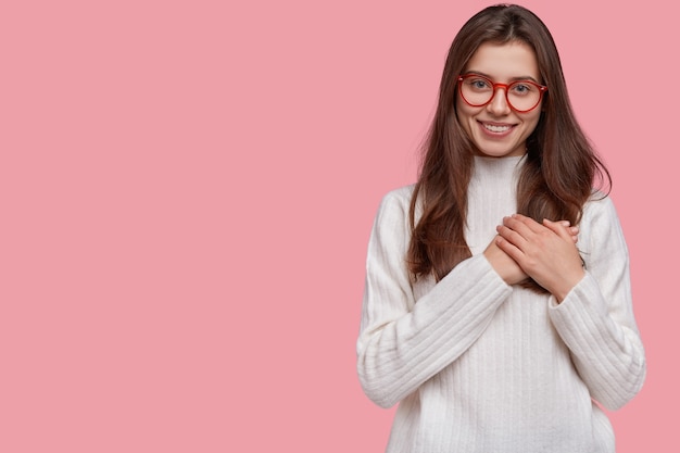 La ragazza tenera gentile preme le mani al cuore in gesto grato e toccato, ha un sorriso piacevole