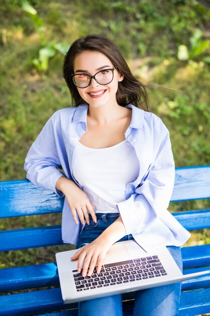 La ragazza tenera in maglietta blu si siede sulla panchina nel parco e usa il suo laptop