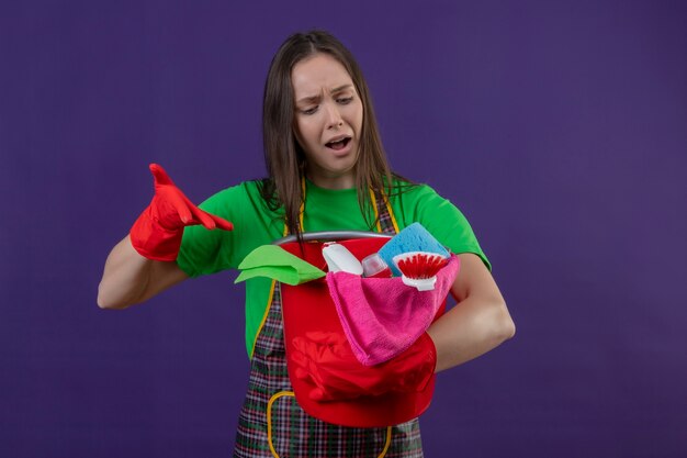 La ragazza triste di pulizia che porta l'uniforme in guanti rossi indica gli strumenti di pulizia sulla sua mano su fondo viola isolato