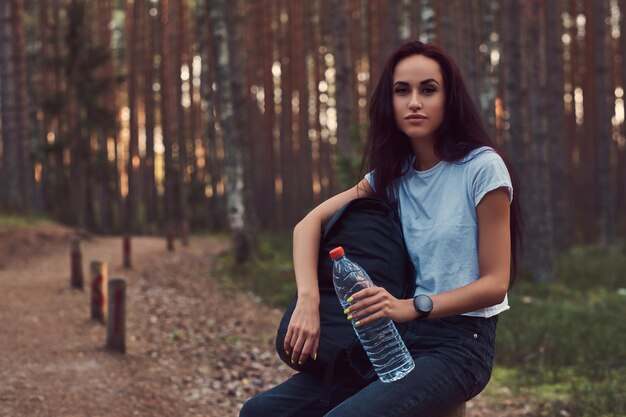 La ragazza turistica dei pantaloni a vita bassa tiene una bottiglia d'acqua, si è fermata a riposare nella bellissima foresta autunnale.