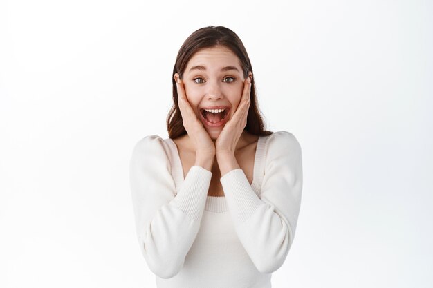 La ragazza urlante sembra sorpresa e stupita, vincendo e provando gioia, tenendosi per mano sul viso e sorridendo, fissando con incredulità le fantastiche grandi notizie, celebrando la vittoria, in piedi sul muro bianco