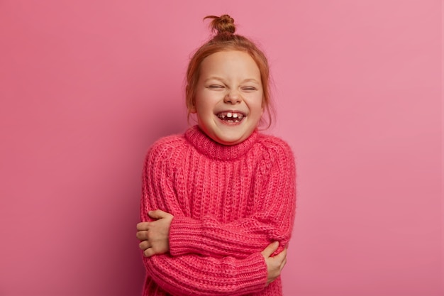La ragazzina zenzero felicissima si coccola, ha un'espressione positiva, indossa un caldo maglione lavorato a maglia, ama il servizio fotografico, esprime emozioni sincere, isolata sul muro rosa. Bambini, intrattenimento