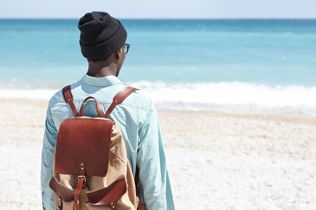 La retrovisione dell'uomo nero europeo che trasporta lo zaino di cuoio marrone che sta soltanto sulla spiaggia del deserto, di fronte al mare azzurrato, è venuto alla spiaggia