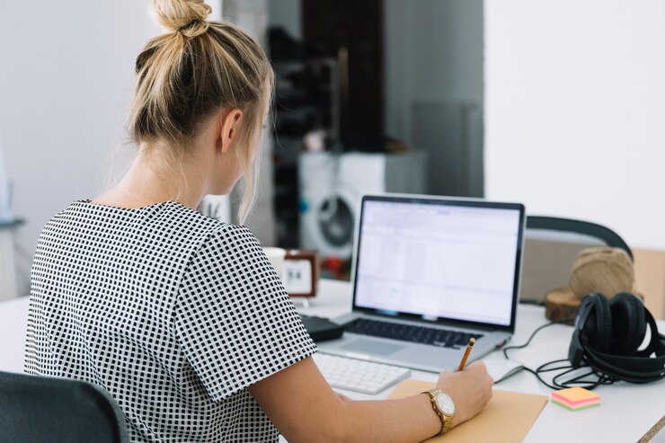 Foto gratuito la retrovisione di una donna di affari scrive le note sopra carta marrone con il computer portatile sullo scrittorio