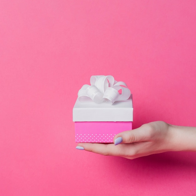 La scatola della tenuta della mano della femmina con l&#39;arco del nastro bianco su fondo rosa