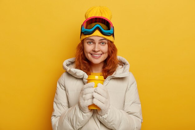 La sciatrice indossa un caldo capospalla invernale, tiene in mano una tazza gialla da asporto con tè caldo, indossa berretto e occhiali da sci, sorride piacevolmente, modella al coperto.