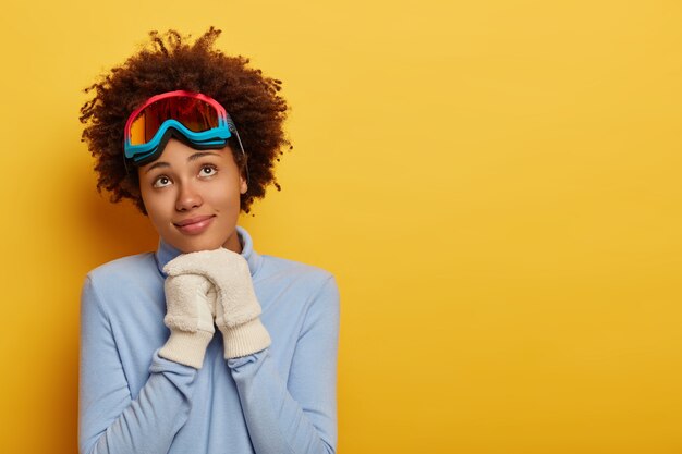 La sciatrice riccia pensierosa indossa un maglione blu caldo e guanti bianchi, ha la maschera da snowboard sulla testa, si erge su sfondo giallo.