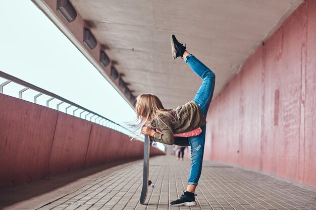 La scolaretta con i capelli biondi vestita con abiti alla moda balla con uno skateboard su un marciapiede del ponte.