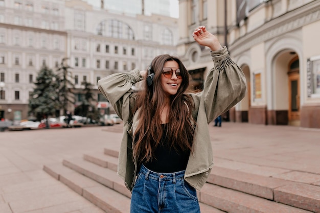 La signora allegra sta ascoltando musica e ballando in città Ragazza abbastanza giovane con lunghi capelli scuri in giacca e jeans con trucco nudo che sorride e socchiude gli occhi mentre agita la testa