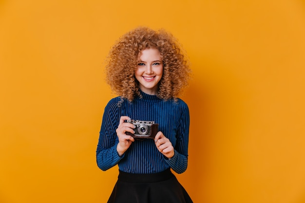 La signora bionda riccia in maglione blu stretto sorride e tiene la retro macchina fotografica sullo spazio giallo.